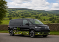 A black Volkswagen Transporter van with 'Dales Tours' branding and a scenic green hillside wrap, parked on a country road with a stone wall and rolling green hills in the background.