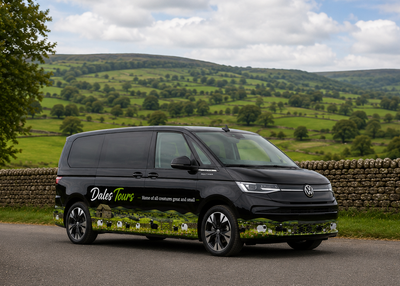 A black Volkswagen Transporter van with 'Dales Tours' branding and a scenic green hillside wrap, parked on a country road with a stone wall and rolling green hills in the background.