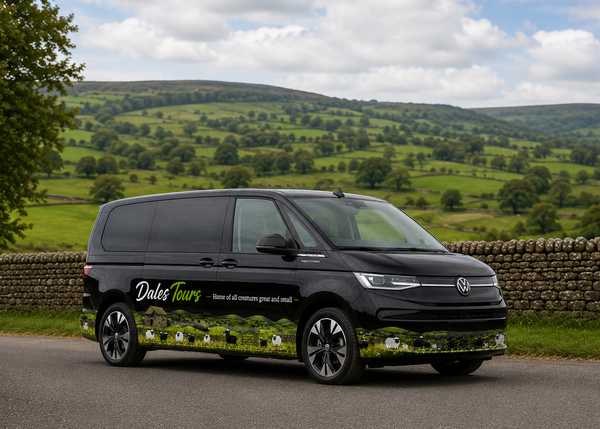 A black Volkswagen Transporter van with 'Dales Tours' branding and a scenic green hillside wrap, parked on a country road with a stone wall and rolling green hills in the background.