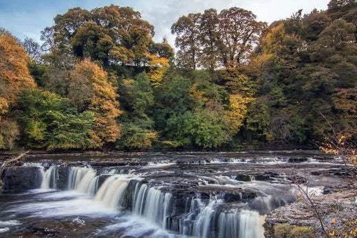 A scenic landscape photograph of a multi-tiered waterfall flowing through a rocky forest bed, surrounded by trees with vibrant green, yellow, and orange autumn foliage under a cloudy blue sky.